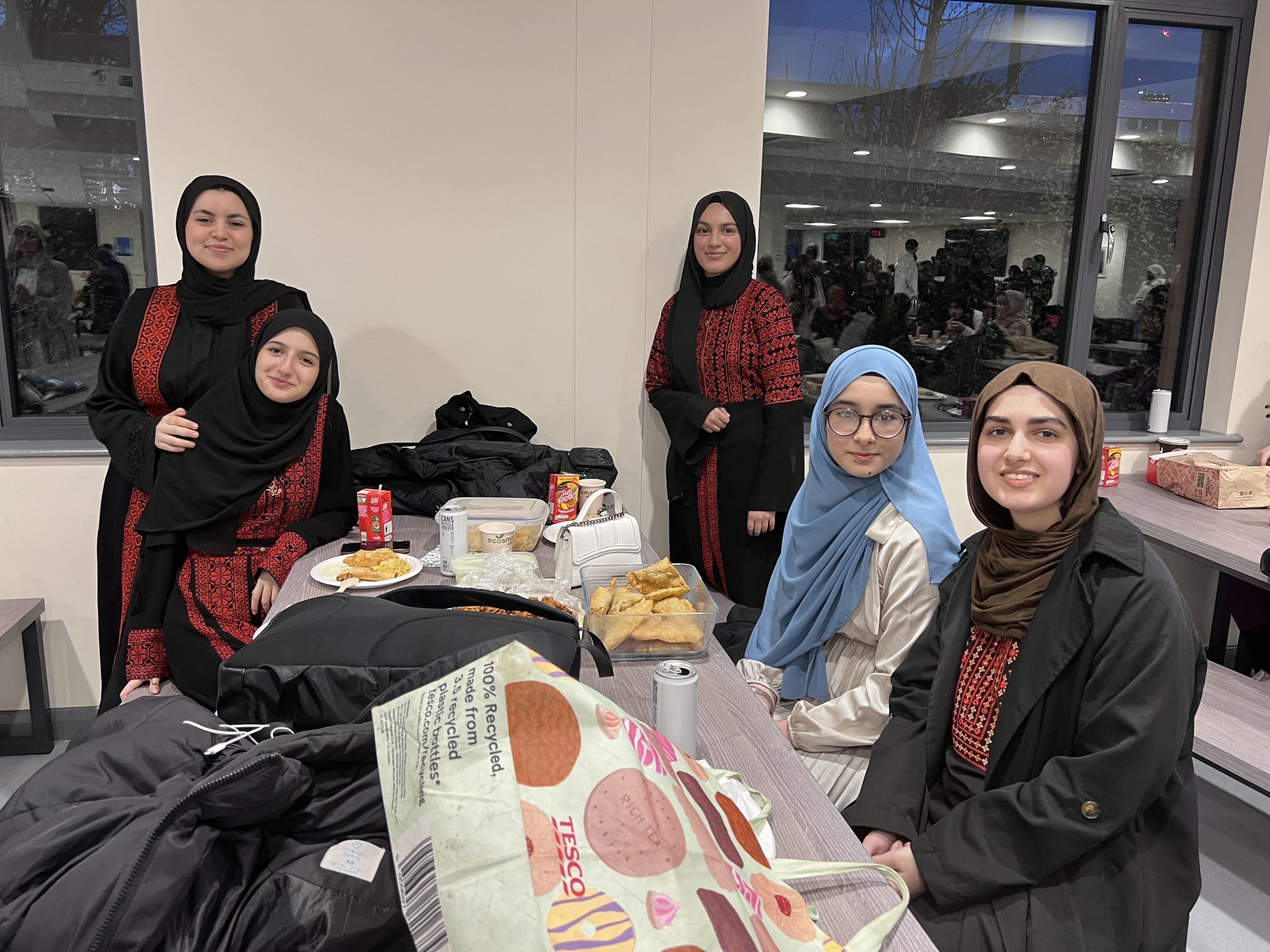 5 Loreto female students, 3 sat by the table with the others stood, smiling for the camera in Loreto cafeteria.