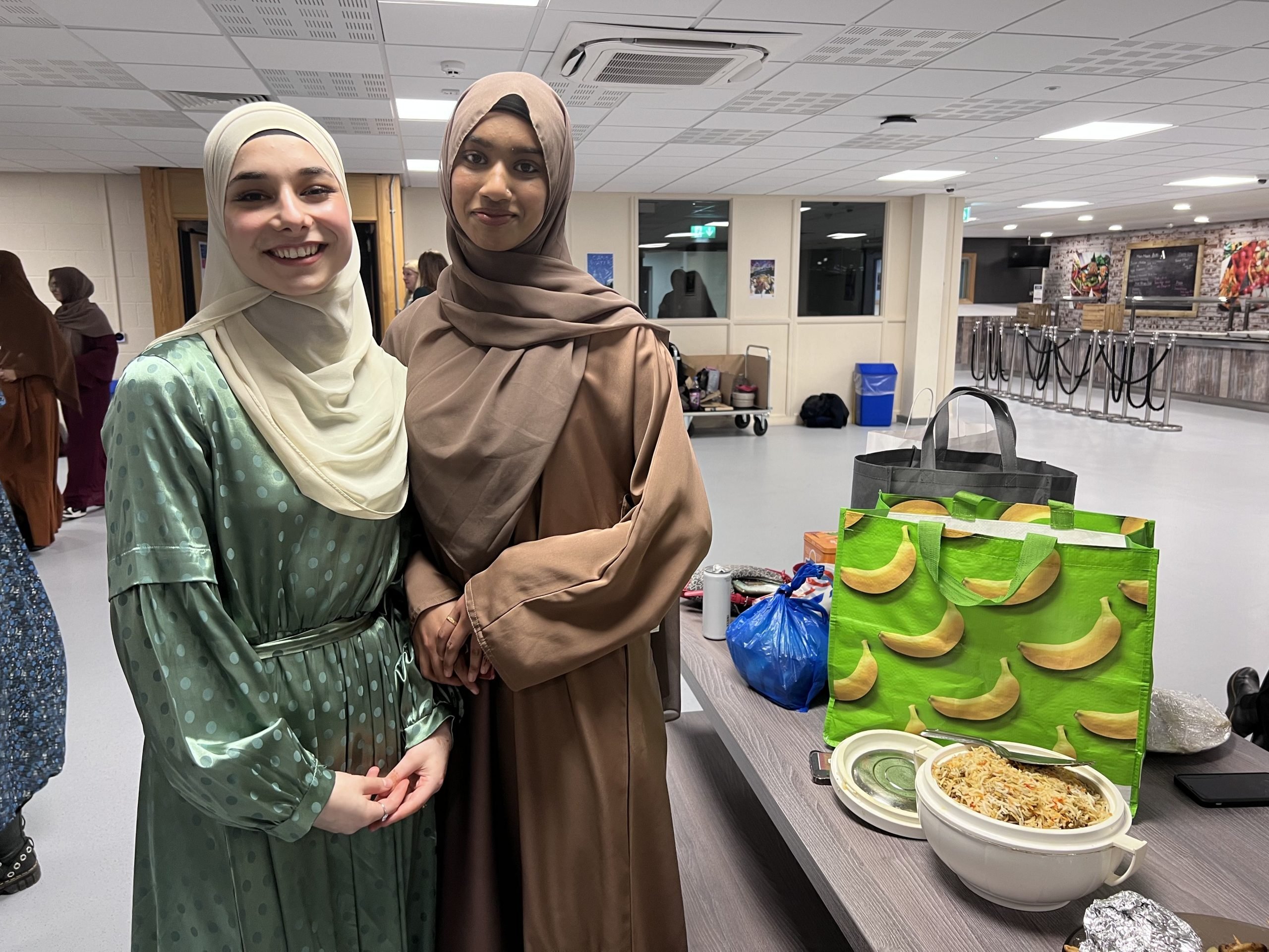 Two Loreto students in hijabs stood smiling by a table with a large container of rice on it.