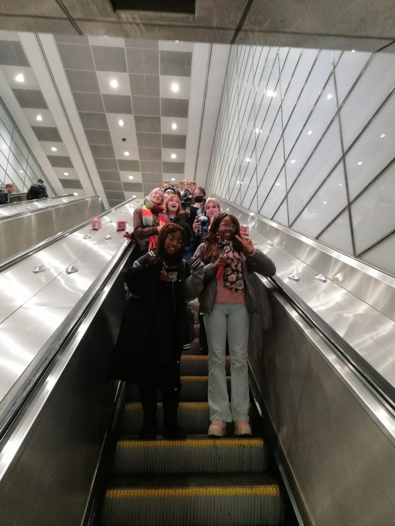 group of students travelling downwards on a London Underground escalator, smiling for the camera.
