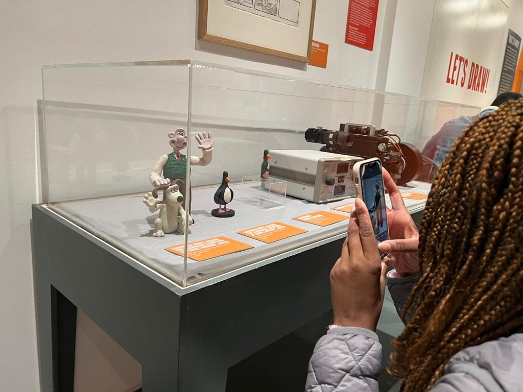 Student taking a picture with their phone, of the Wallace and Gromit figures in a box display cabinet.