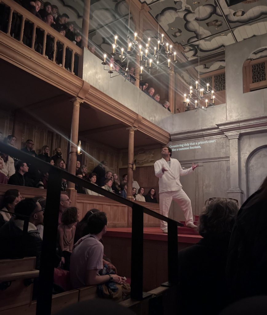 Audiences sat in the stalls and balcony seats watch an actor dressed in a white suit performing.