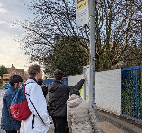A group of students waiting at a bus stop reading the bus timetable with their teacher.