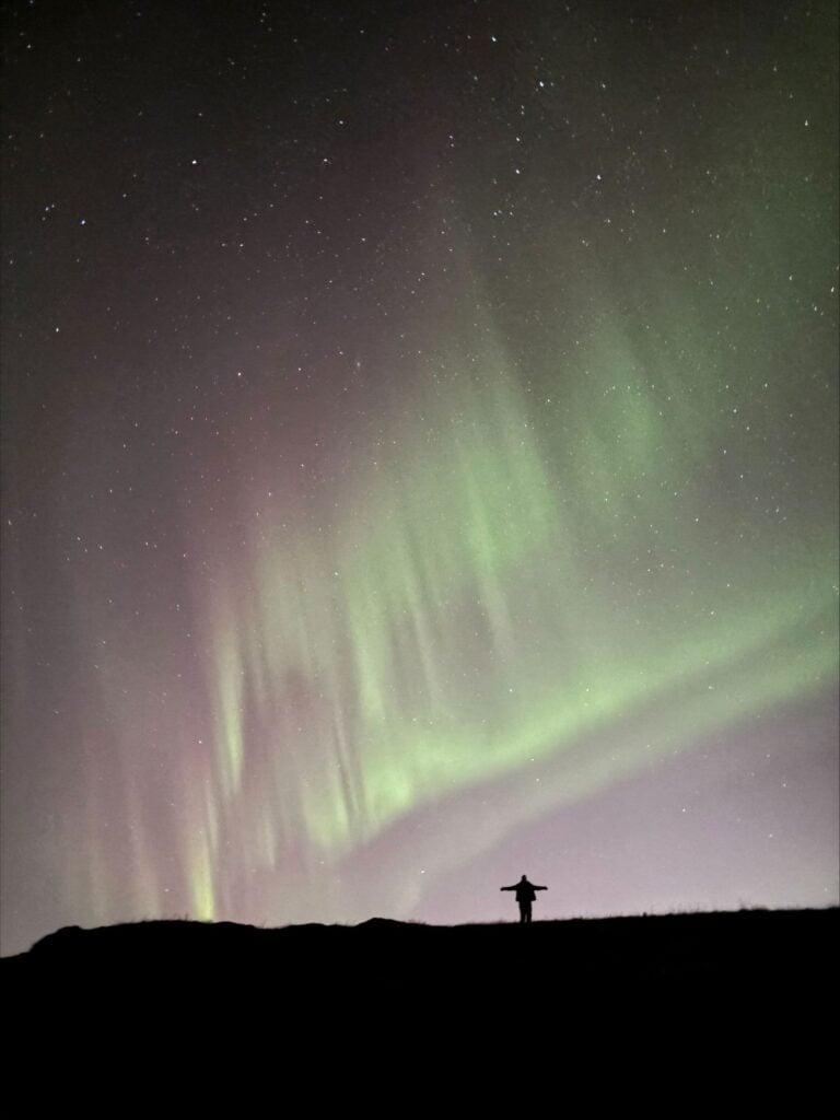 Silhouetted figure beneath green and purple Northern Lights in the night sky.