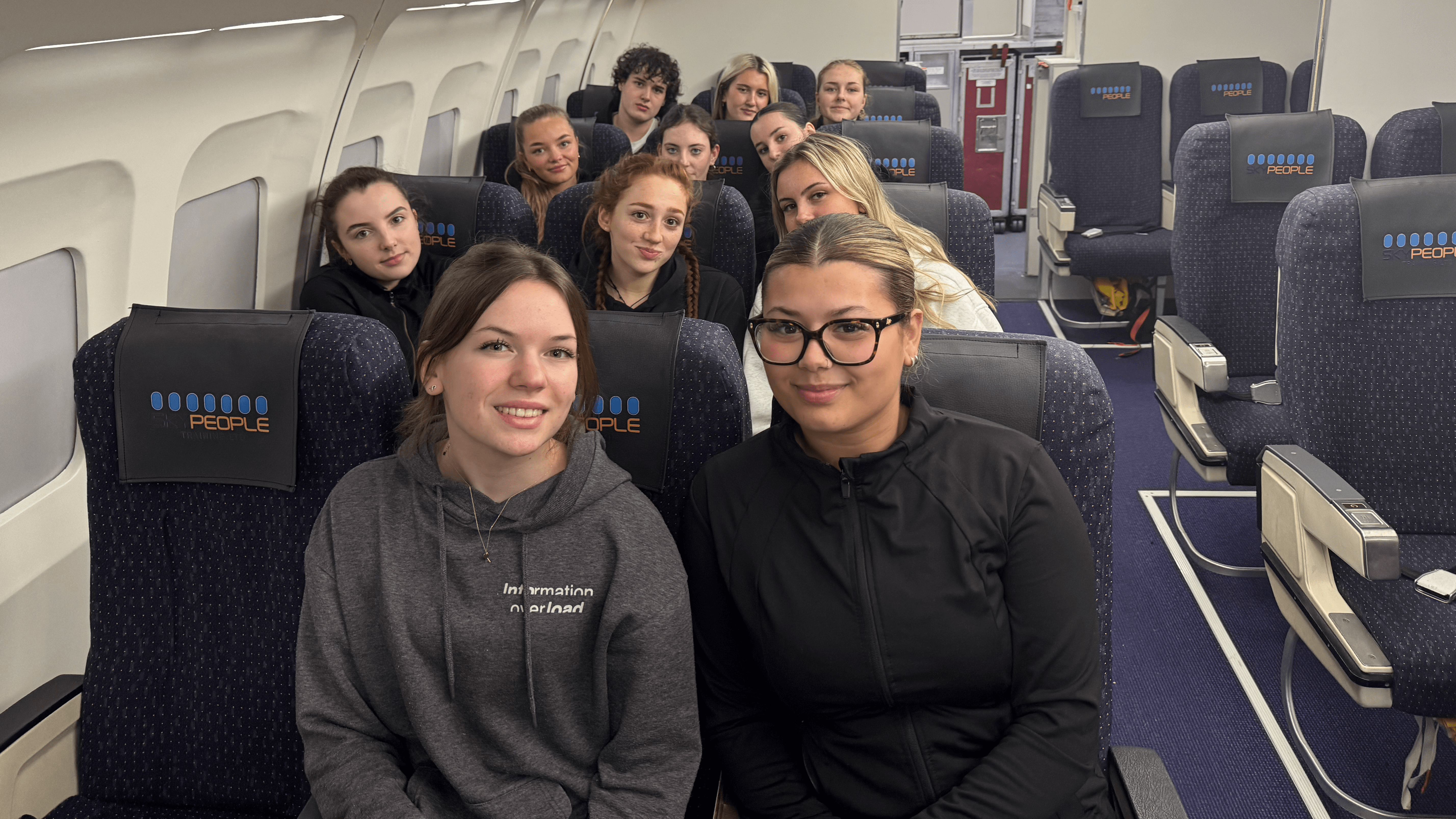 Students seated inside a realistic aircraft cabin training facility at Skypeople, with rows of airline seats, overhead lockers, and cabin equipment visible.