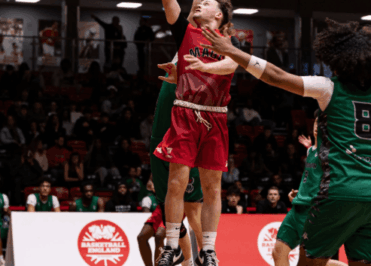 A basketball player in red leaps toward the hoop for a layup during the Elite Academy Basketball League Final, while a Loreto College player in green attempts to block the shot.