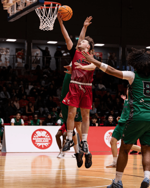 A basketball player in red leaps toward the hoop for a layup during the Elite Academy Basketball League Final, while a Loreto College player in green attempts to block the shot.