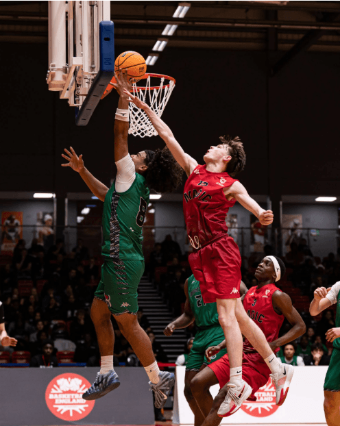 A Loreto College basketball player in green jumps to defend a shot at the rim during the Elite Academy Basketball League Final against an opposing player in red.