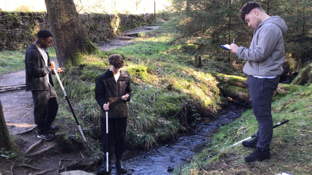 Geography students measuring river depth and recording data during River Bollin fieldwork.