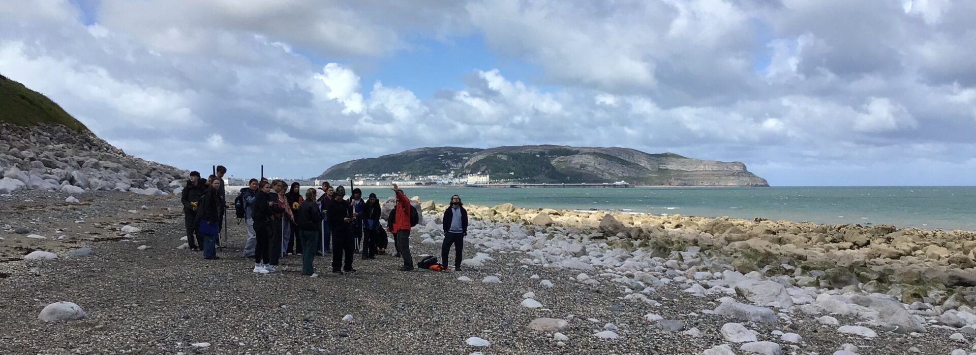 A group of Loreto Geography students standing on a rocky beach in Llandudno with the Great Orme headland visible across the bay.