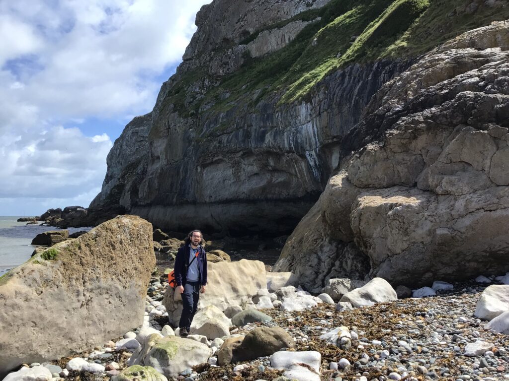 A Loreto student standing among large coastal boulders beneath steep limestone cliffs at the Great Orme in Llandudno.
